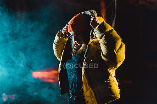 Stylish young woman standing at night and putting on hood of yellow jacket. — Stock Photo