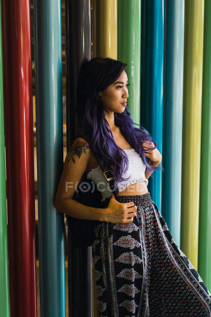 Jovem mulher com cabelo roxo encostado em colunas coloridas e olhando para longe . — Fotografia de Stock