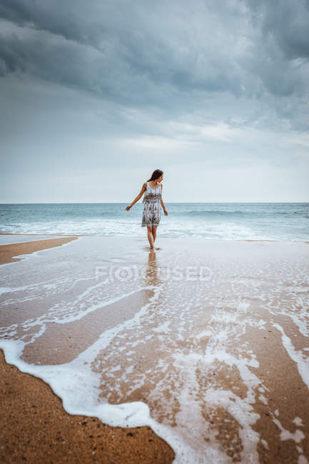 Jeune femme en robe marchant dans les eaux peu profondes de l'océan vague sous un ciel sombre
. — Photo de stock