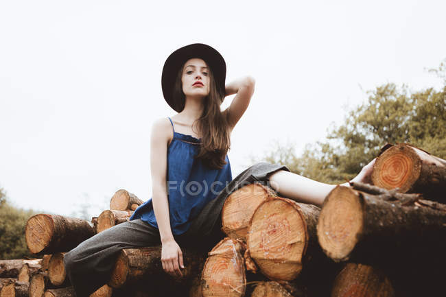 Sensuale bruna donna in cappello seduta sul mucchio di tronchi — Foto stock