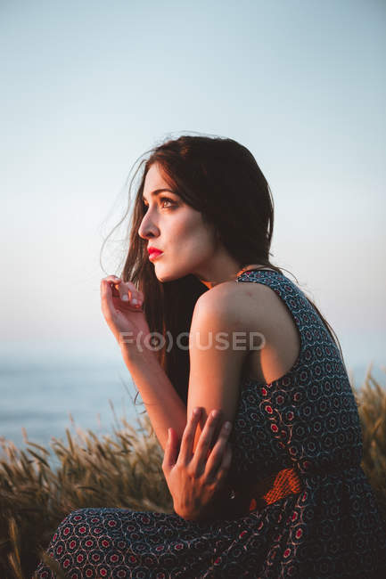 Brunette girl in dress sitting in field on shoreline — Stock Photo