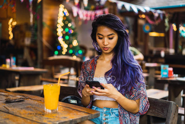 Junge Frau sitzt mit Orangensaft am Cafétisch und surft im Smartphone. — Stockfoto