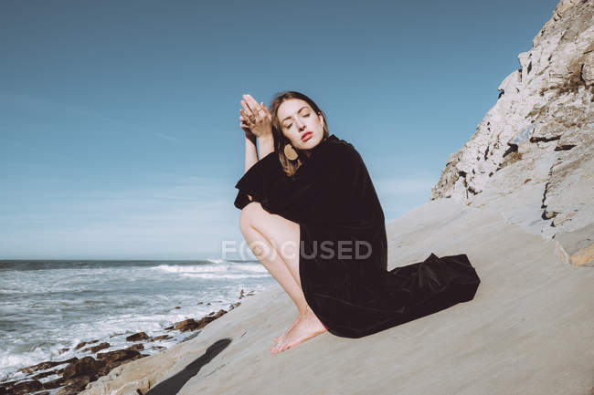 Jovem morena de casaco preto sentada na costa com os olhos fechados — Fotografia de Stock