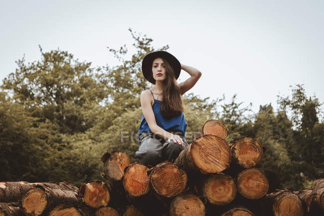 Brunette woman in hat sitting on log pile and looking at camera — Stock Photo