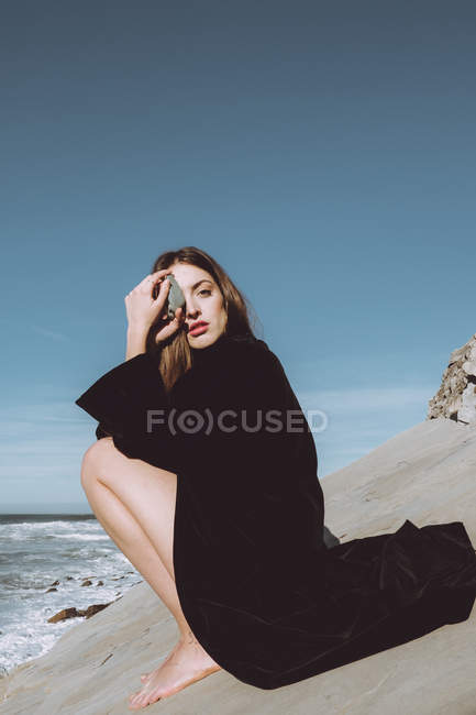 Брюнетка в черном пальто сидит на берегу и держит камешек перед глазами — стоковое фото