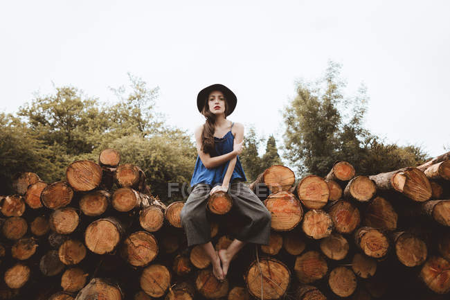 Stylish brunette woman in hat sitting on log pile — Stock Photo