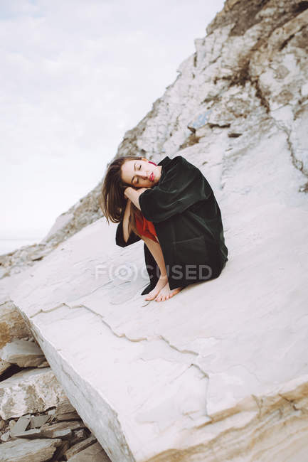 Stylish brunette sitting on shore rocks with closed eyes — Stock Photo