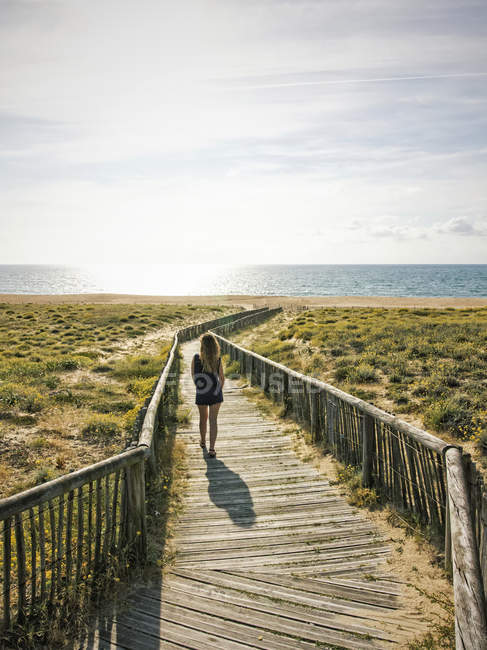 Rückansicht einer Frau, die an der Strandpromenade steht, die an einem sonnigen Tag zum Strand am Meer führt. — Stockfoto