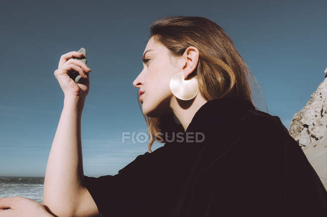 Junges Mädchen in schwarzem Mantel sitzt an der Küste und blickt auf Kieselsteine in der Hand — Stockfoto