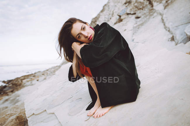 Stylish brunette sitting on shore rock and looking at camera — Stock Photo