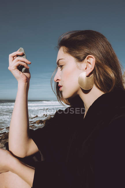 Jovem de casaco preto sentado na costa e com seixo na mão — Fotografia de Stock