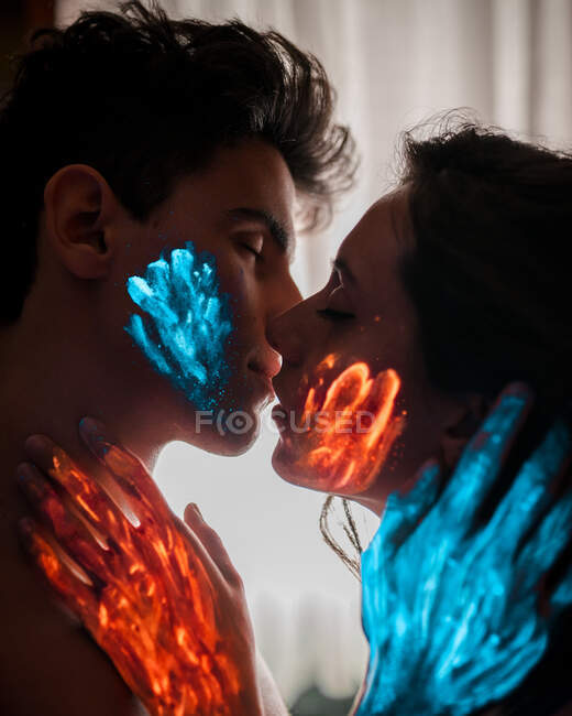 Молодая пара с оранжевой и голубой сияющей краской целуется с закрытыми глазами. — стоковое фото