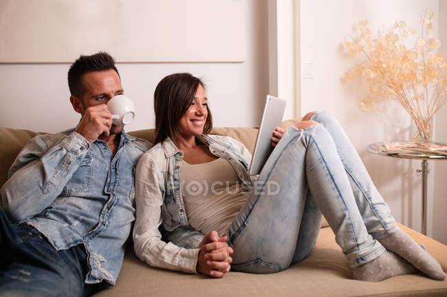 Jeune couple joyeux assis sur le canapé ensemble et en utilisant la tablette . — Photo de stock