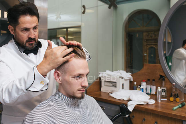 Barbiere styling capelli di giovane cliente seduto in salone . — Foto stock