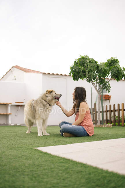 Вид сбоку на молодую женщину, сидящую на газоне на заднем дворе и гладящую собаку. — стоковое фото