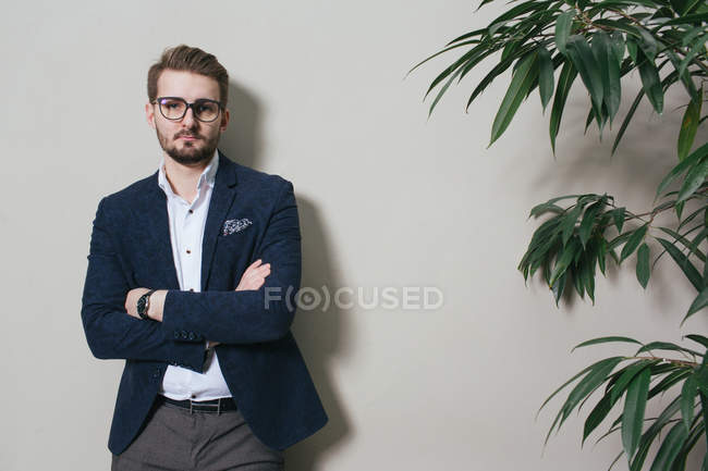 Junger Mann in Anzug und Brille steht mit verschränkten Armen an Topfpflanze. — Stockfoto