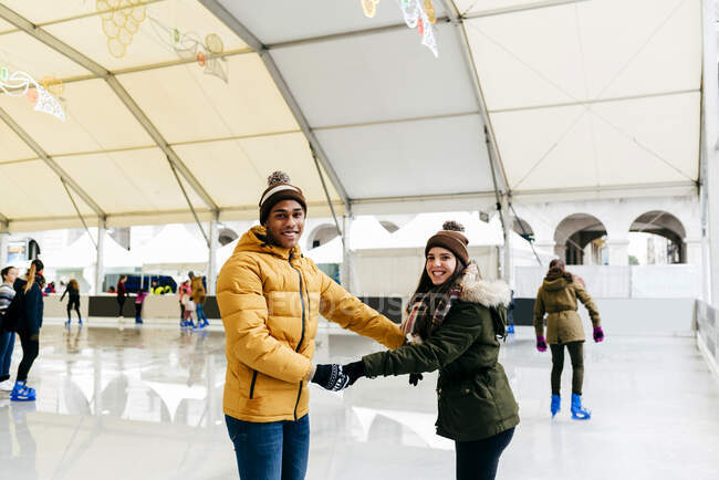 Fröhliches junges multiethnisches Paar hält Händchen, während es auf Eisbahn reitet. — Stockfoto