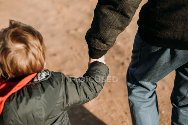 Cultivo disparado desde la espalda del hombre sosteniendo la mano de un niño pequeño caminando bajo la luz del sol. - foto de stock