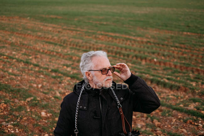 Älterer ernsthafter Mann mit Sonnenbrille und schwarzer Jacke blickt vertrauensvoll auf den Hintergrund des Feldes. — Stockfoto