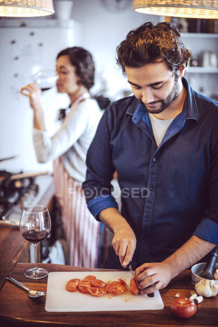 Мужчина режет помидоры на кухне над женщиной, пьющей вино — стоковое фото