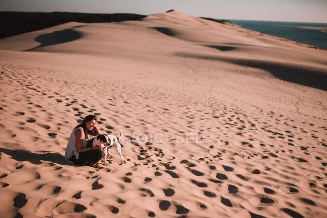 Femme gaie avec chien assis sur le sable dans la journée ensoleillée . — Photo de stock