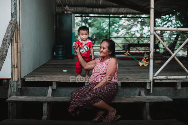 LAOS, 4000 ILHAS ÁREA: Mulher sênior sentada em degraus de casa de madeira e segurando a mão da criança . — Fotografia de Stock