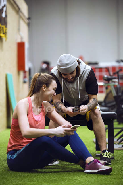 Cheerful sporty man and woman sitting in gym and browsing smartphones. — Stock Photo