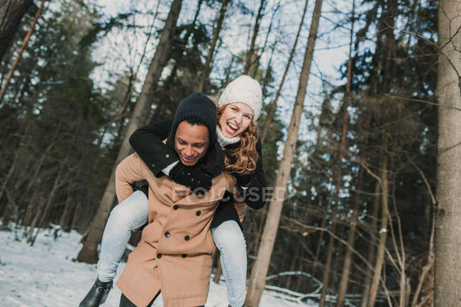 Cheerful man carrying girlfriend on back in winter forest. — Stock Photo