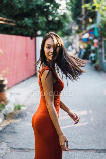 Cheerful woman in red dress walking on street and looking over shoulder at camera — Stock Photo