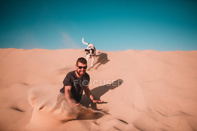 Hombre alegre en gafas de sol deslizándose sobre duna de arena con perro en día soleado
. - foto de stock