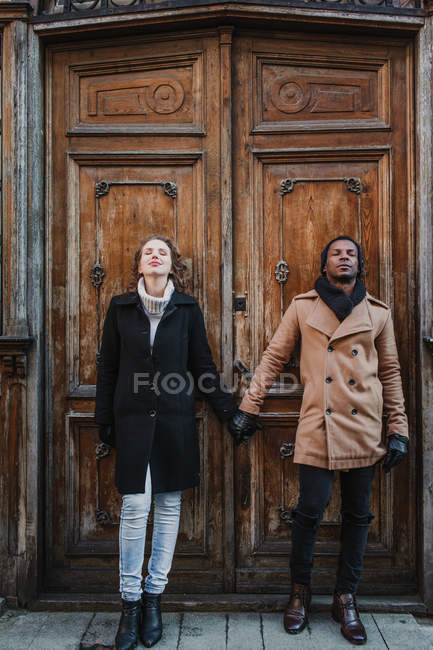 Paar in warmer Kleidung stehend und Händchen haltend mit geschlossenen Augen an grunziger alter Holztür. — Stockfoto