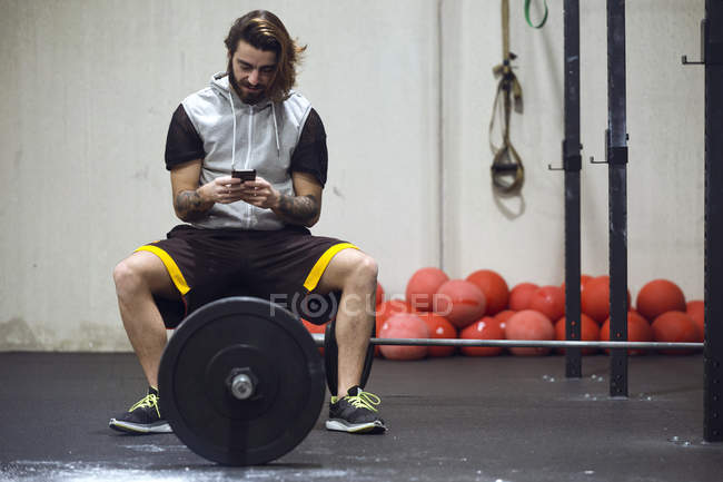 Sportsman sitting on barbell and browsing smartphone in gym. — Stock Photo