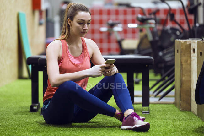 Frau sitzt im Fitnessstudio auf dem Boden und surft mit Smartphone — Stockfoto