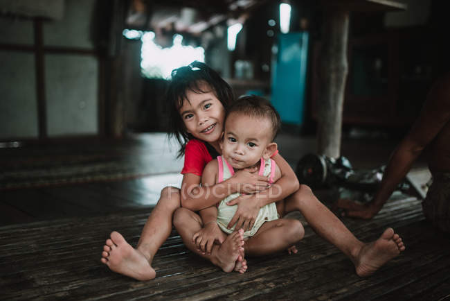 Laos, 4000 inseln: lächelndes mädchen umarmt kleinkind und blickt in kamera, während sie auf holzboden sitzt. — Stockfoto