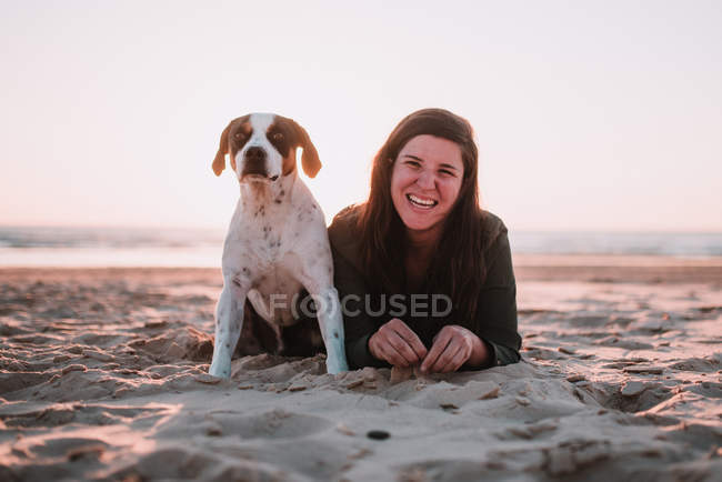 Портрет веселої жінки з маленькою собакою, що лежить на піску і дивиться на камеру — стокове фото