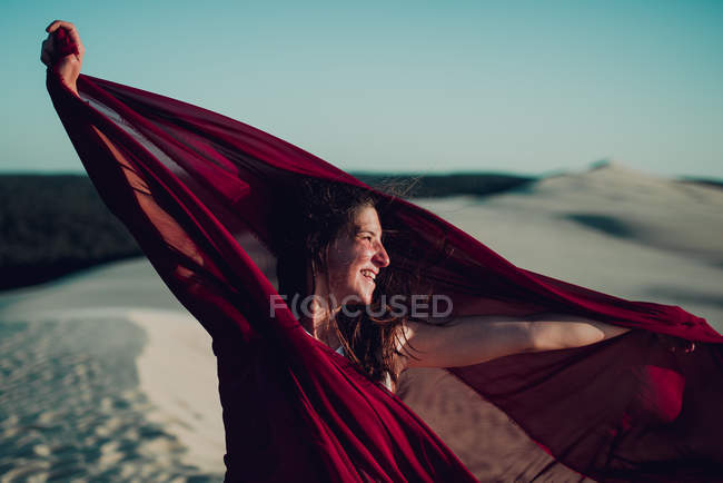 Vista laterale di donna allegra in tessuto rosso in posa su dune di sabbia — Foto stock