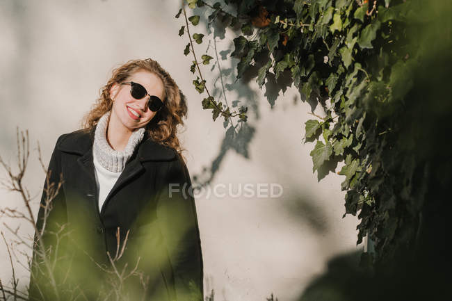 Весела жінка в теплому одязі і сонцезахисних окулярах стоїть біля стіни і куща . — стокове фото