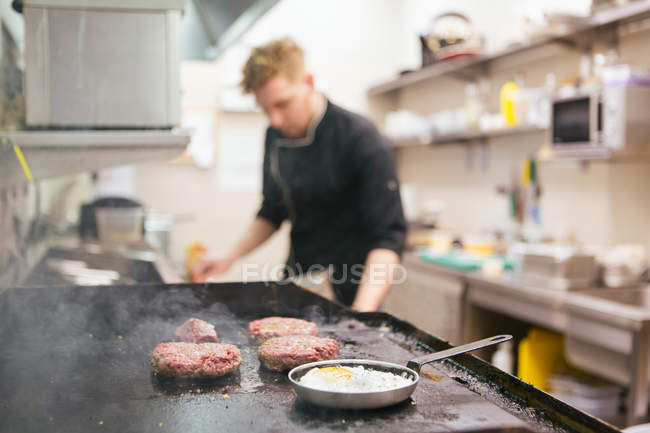 Unrecognizable cook standing on kitchen of restaurant while patties and eggs frying on stove — Stock Photo