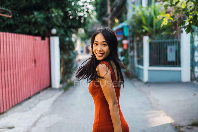 Cheerful woman in red dress looking over shoulder at camera at street scene — Stock Photo