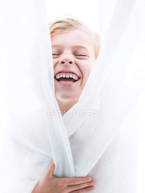 Cheerful young boy wrapping in curtains and laughing — Stock Photo