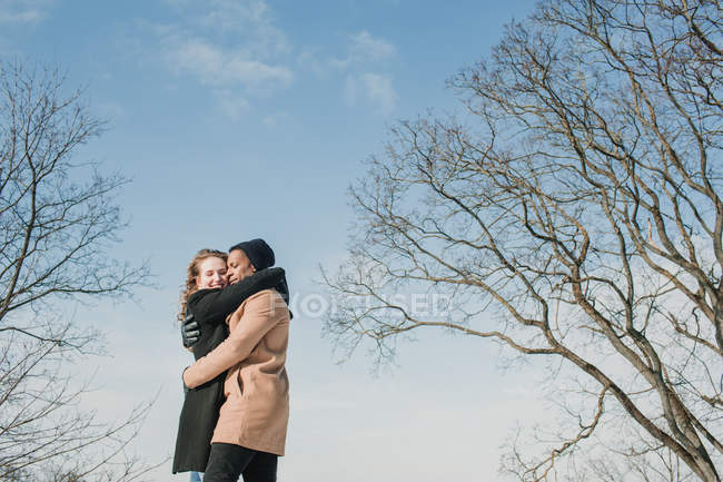 Fröhliches Paar in warmen Kleidern, das sich bei sonnigem Wetter an blattlosen Bäumen umarmt. — Stockfoto