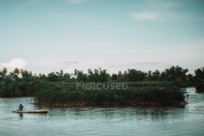 Vista distante dell'uomo seduto su una barca e che lo guida su un ampio fiume vicino a una piccola isola . — Foto stock
