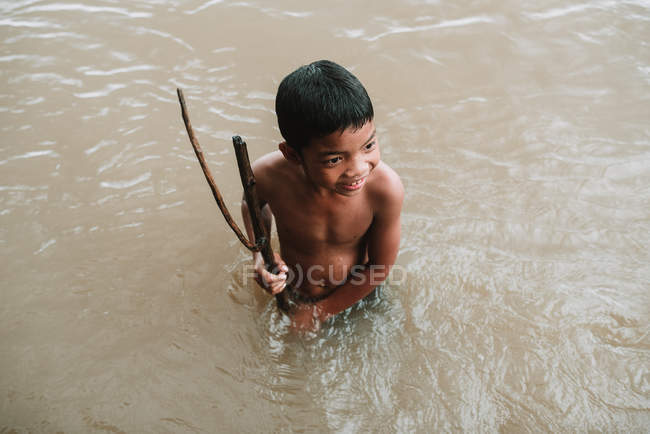 LAOS, 4000 ÎLES : Garçon avec des bâtons debout dans une rivière sale — Photo de stock