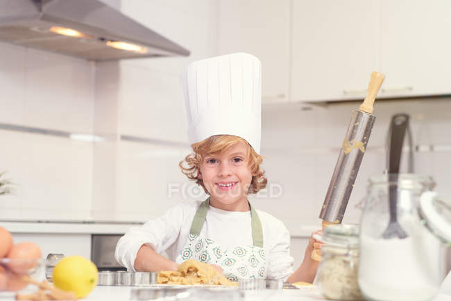 Mignon gai petit garçon rouler pétrir sur cuisine à la maison en regardant caméra — Photo de stock