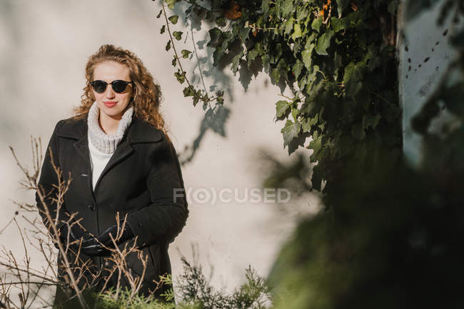 Cheerful young woman in sunglasses posing by ivy-embracing wall — Stock Photo