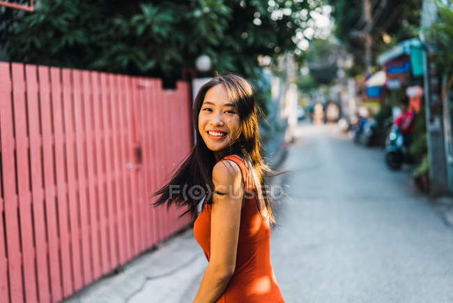 Femme gaie en robe rouge posant sur la rue et regardant par-dessus l'épaule à la caméra — Photo de stock