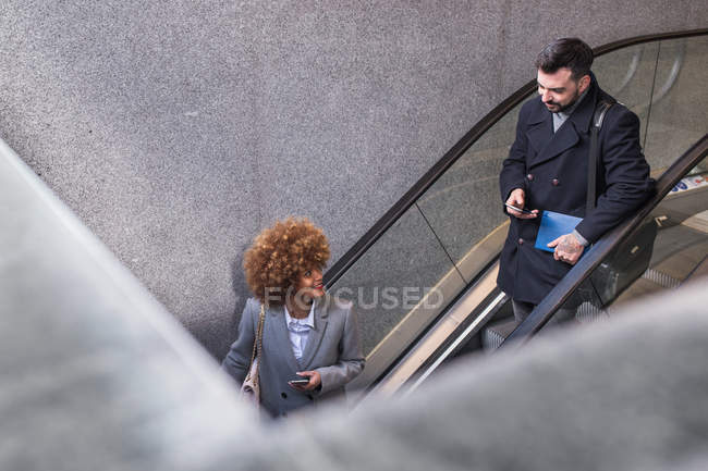 Стильная пара с телефонами в руках, смотрящими друг на друга на эскалаторе — стоковое фото