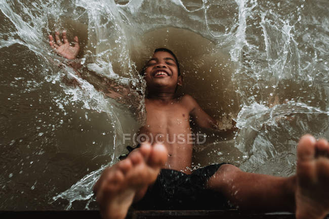 LAOS, 4000 ISLAS ÁREA: Niño sin camisa en pantalones cortos riendo mientras cae en el agua del río sucio . - foto de stock