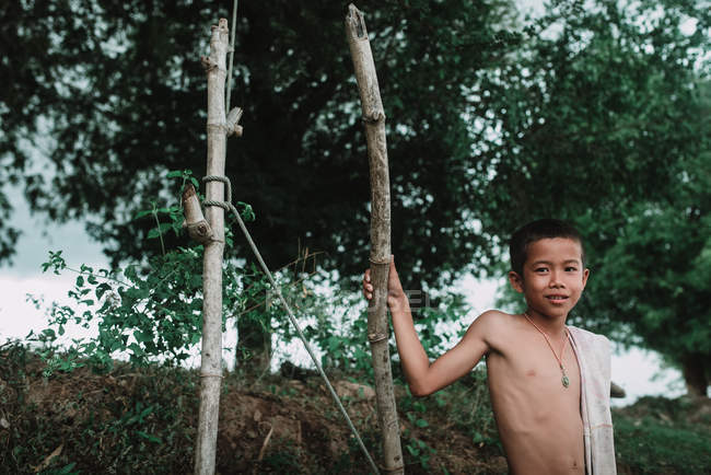 LAOS, 4000 ISLAS ÁREA: Niño sin camisa de pie en la naturaleza cerca de dos postes de madera y sonriendo . - foto de stock