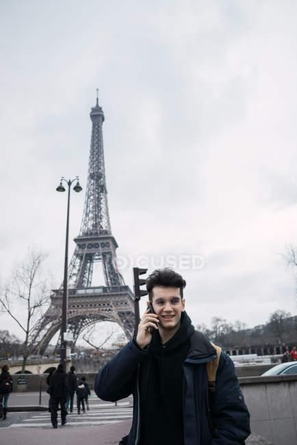 Улыбающийся молодой человек разговаривает по смартфону на фоне Эйфелевой башни . — стоковое фото
